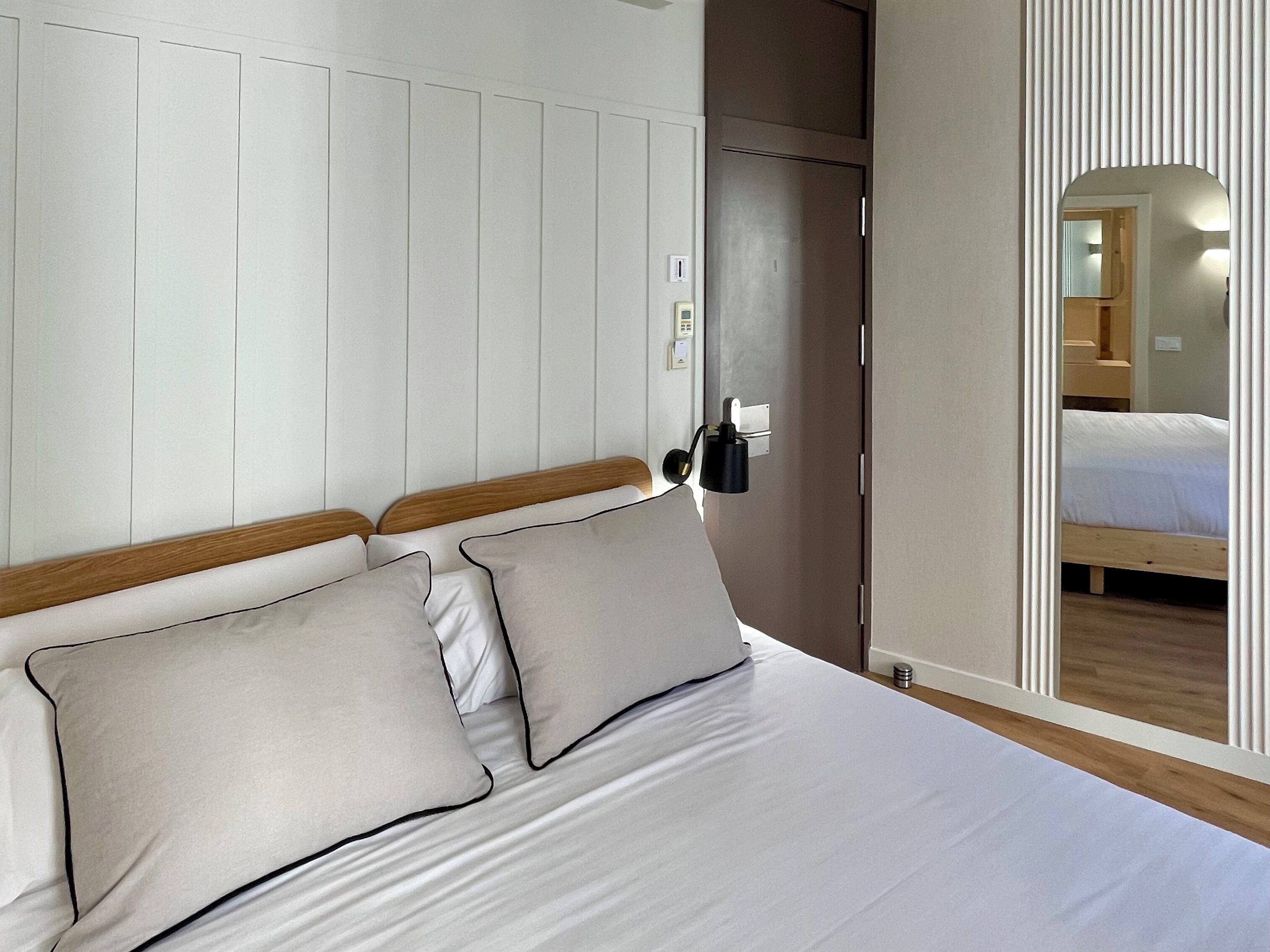 A bedroom with a bed, two pillows, a nightstand, a wall-mounted lamp, and a door leading to another room.