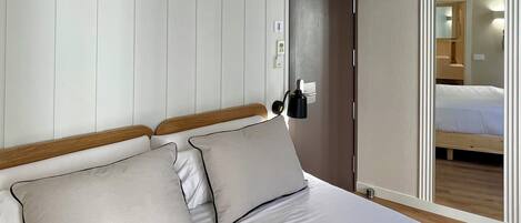 A bedroom with a bed, two pillows, a nightstand, a wall-mounted lamp, and a door leading to another room.