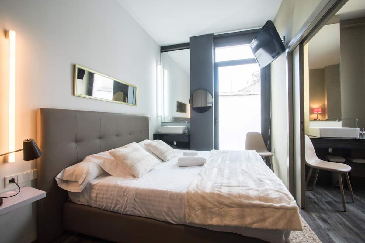 A modern hotel room with a large bed, bedside tables, a flat-screen TV, a mirror, and a bathroom with a sink and chair.