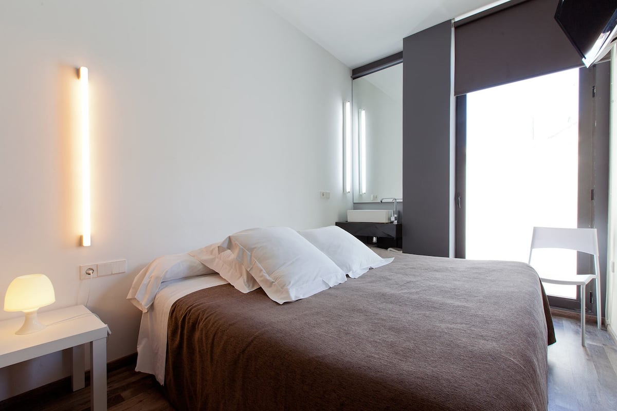 A modern bedroom with a large bed, a bedside table with a lamp, a chair, and a large window.