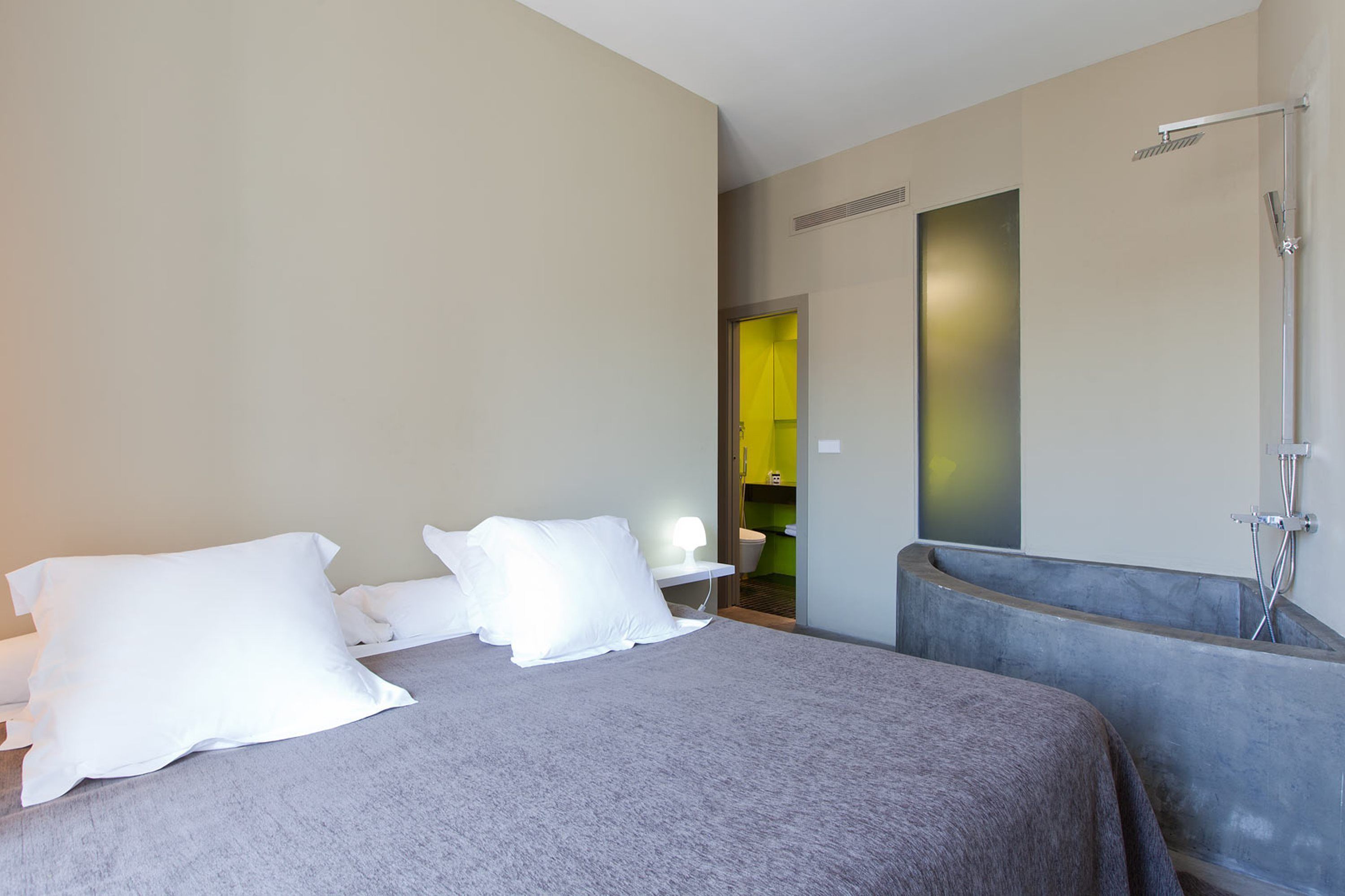 Una habitación de hotel moderna con una cama grande, zona de ducha y una pequeña mesa con lámpara.