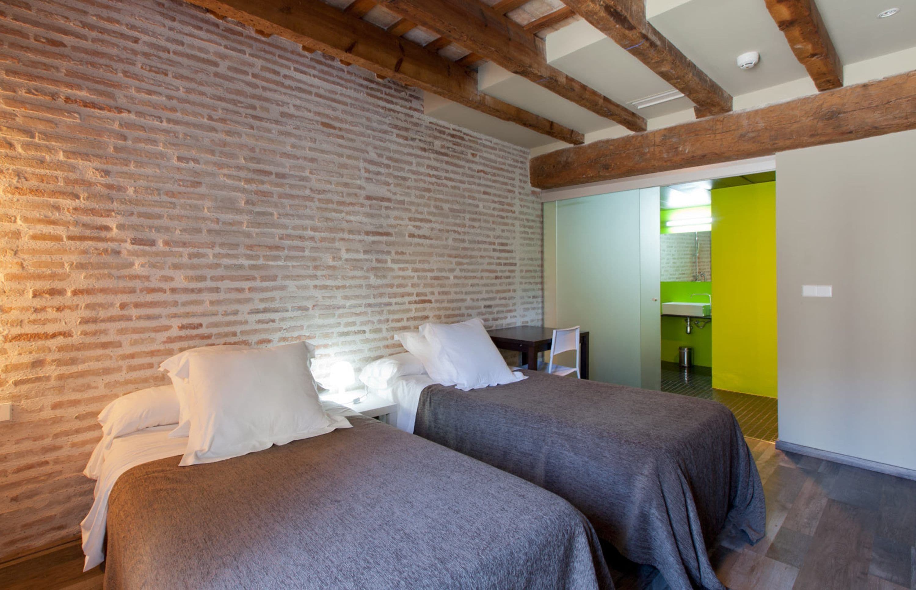 Ein Zimmer mit zwei Betten, einer Ziegelwand, einer Holzbalkendecke und einer hellgrünen Wand im Hintergrund.