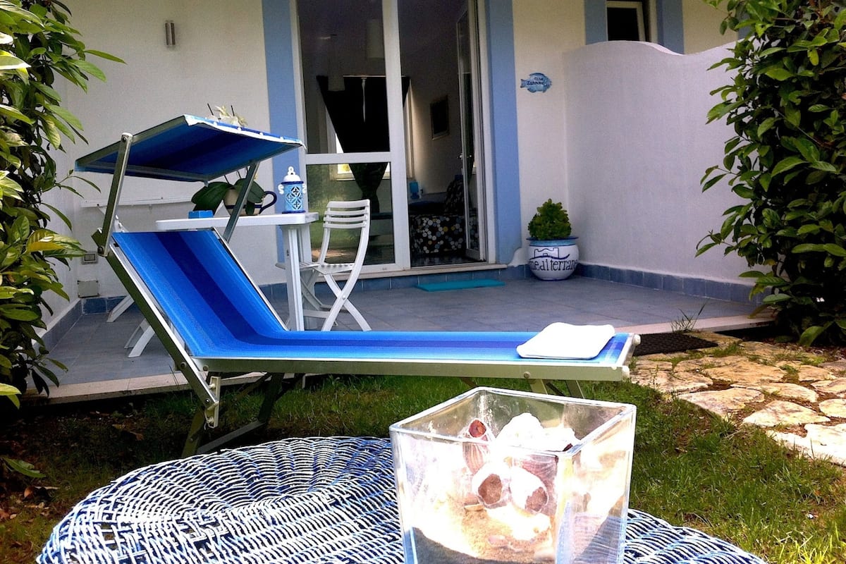 A blue lounge chair with a matching umbrella, a small table with a white cloth, and a potted plant on a patio with a glass door leading to an interior space.