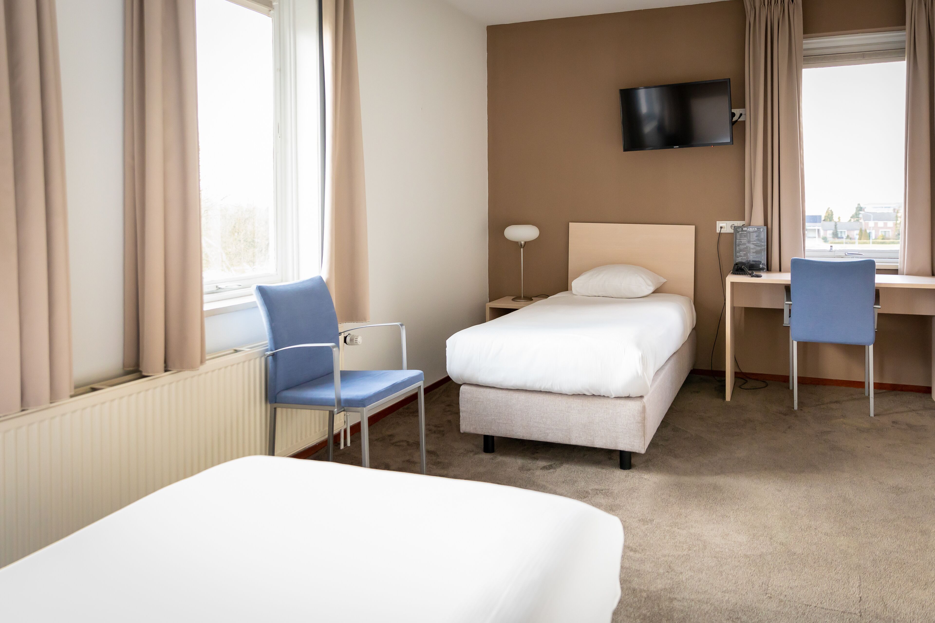 Une chambre d’hôtel comprenant un lit, un bureau avec une chaise, un téléviseur fixé au mur et une fenêtre avec des rideaux.