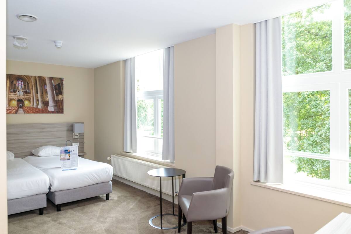 Habitación de hotel con cama, una mesita, una silla y ventanas amplias con vistas a zonas verdes.
