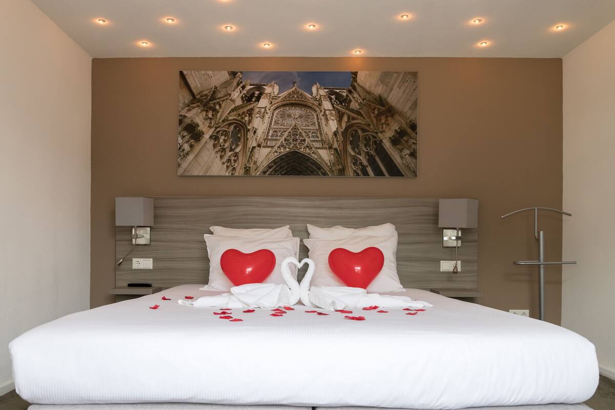Una habitación de hotel con una cama grande, un cabecero con un panel decorativo, una obra de arte colgada en la pared que representa una catedral, y una mesita de noche con una lámpara.