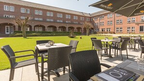 Ein Außenbereich mit einer Terrasse, Korbmöbeln und einem Menüständer, vor einem Ziegelgebäude mit mehreren Fenstern und einem gepflegten Rasen.