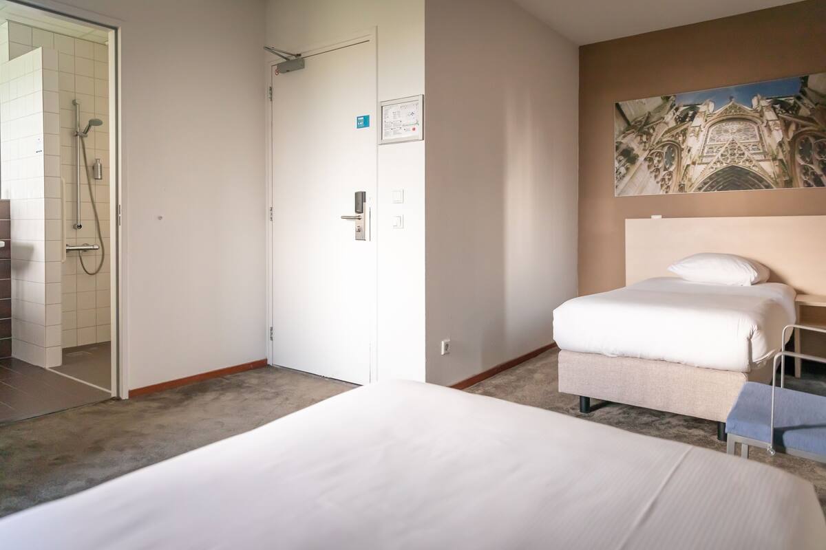 Una habitación de hotel con cama, un pequeño banco, zona de ducha y un cuadro en la pared.