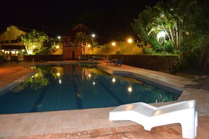 A swimming pool with a lounge chair in the foreground, surrounded by a landscaped garden and a building with illuminated windows. - Hotel Lago das Pedras (Arapongas)