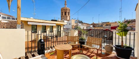 A rooftop terrace with a table, chairs, potted plants, and a view of a tower in the background.