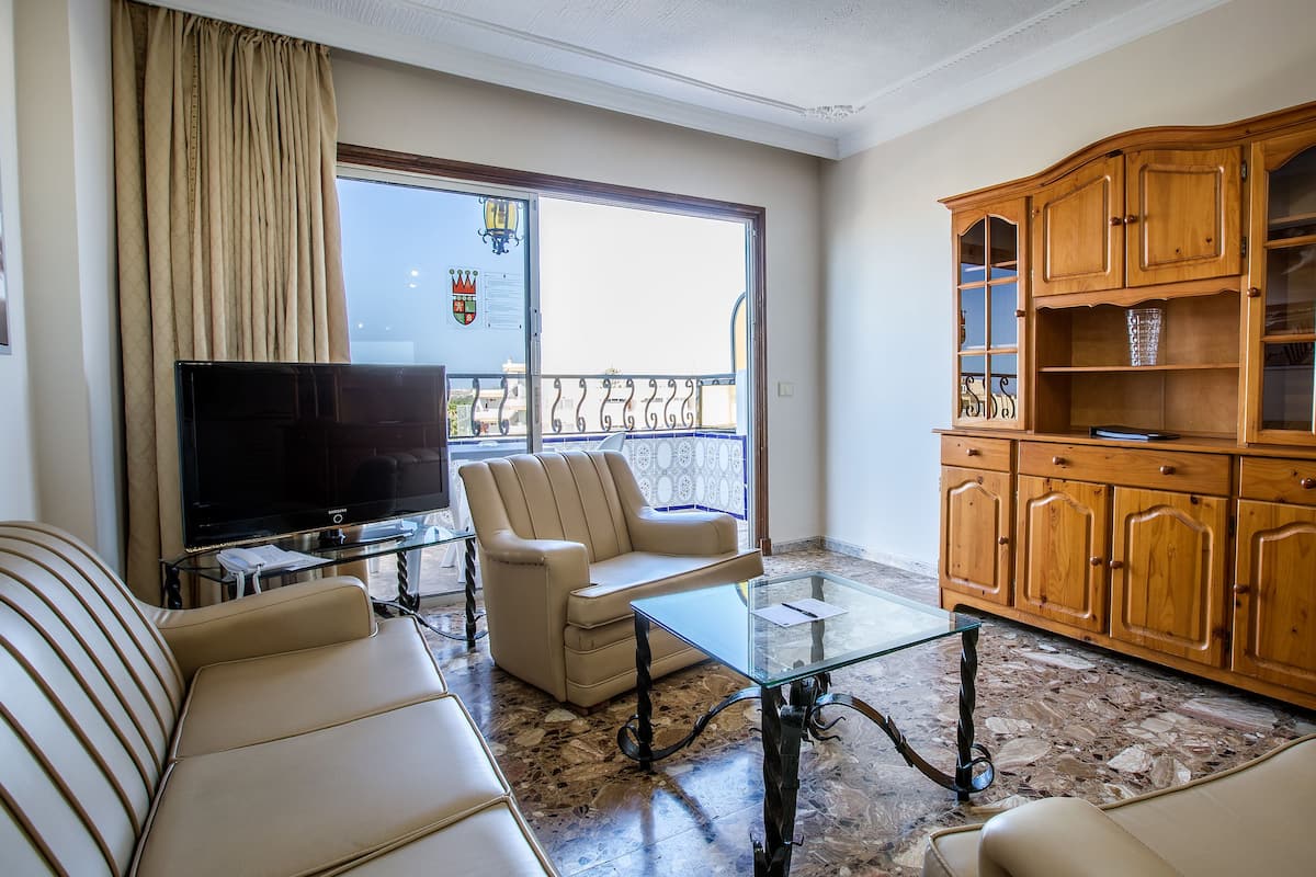 Ein Wohnzimmer mit Sofa, Sessel, Glastisch, Fernseher und Holzschrank.