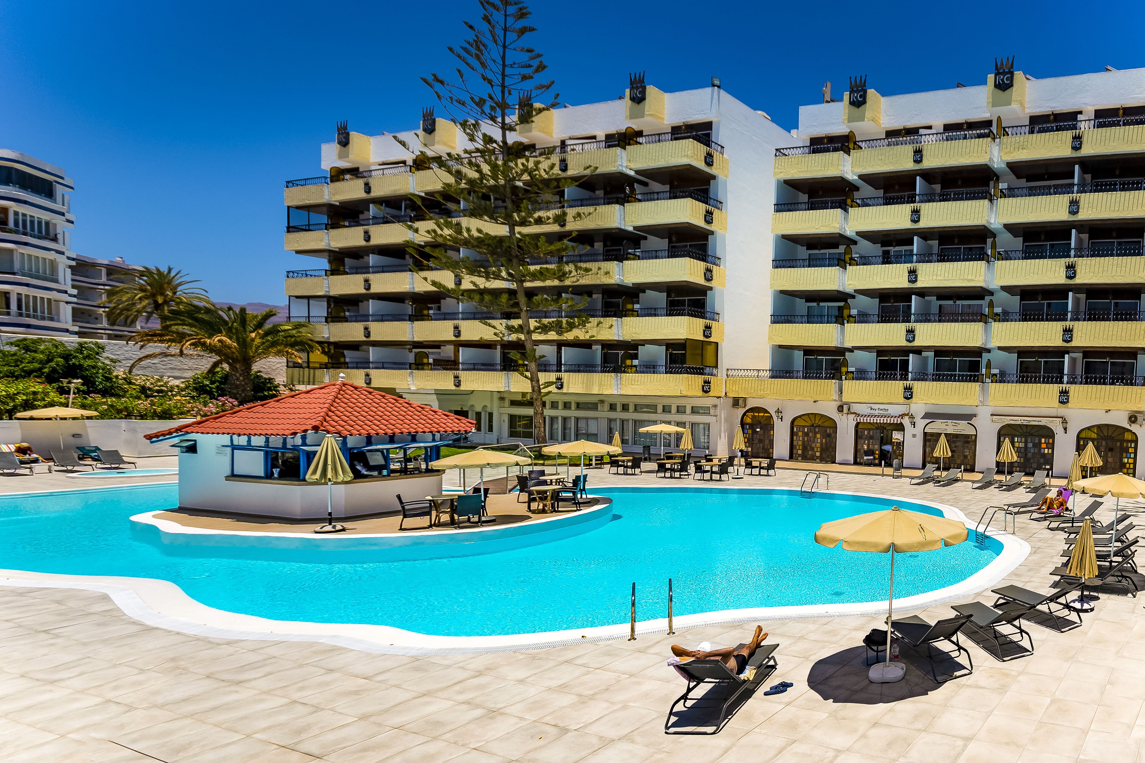 A hotel with a large swimming pool, sun loungers, and a bar area.