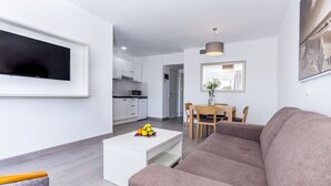 Una sala de estar moderna con un televisor de pantalla plana, una mesa de centro blanca con fruta, un sofá marrón y un comedor con una mesa y sillas de madera.