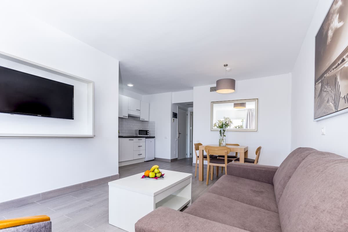 Ein modernes Wohnzimmer mit Flachbildfernseher, einem weißen Couchtisch mit Obst, einer braunen Couch und einem Essbereich mit Holztisch und Stühlen.