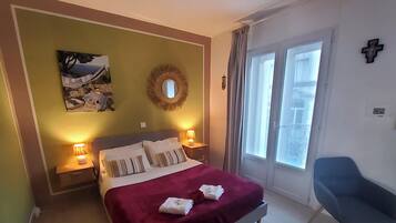 Ein Hotelzimmer mit einem Bett, zwei Nachttischlampen, einem Fenster mit Vorhängen, einem blauen Sessel und einem an der Wand befestigten Kruzifix.