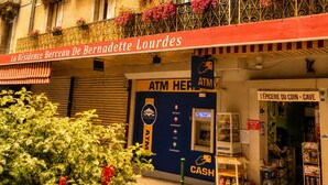 Un immeuble doté de balcons ornés de fleurs, une enseigne indiquant « La Résidence Berceau De Bernadette Lourdes », et un distributeur automatique de billets au rez-de-chaussée.