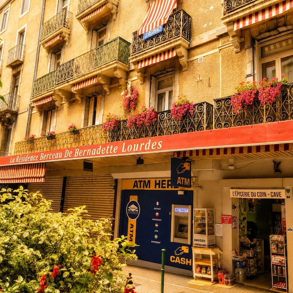 A building with balconies adorned with flowers, a sign for 'La Résidence Berceau De Bernadette Lourdes,' and an ATM on the ground floor.