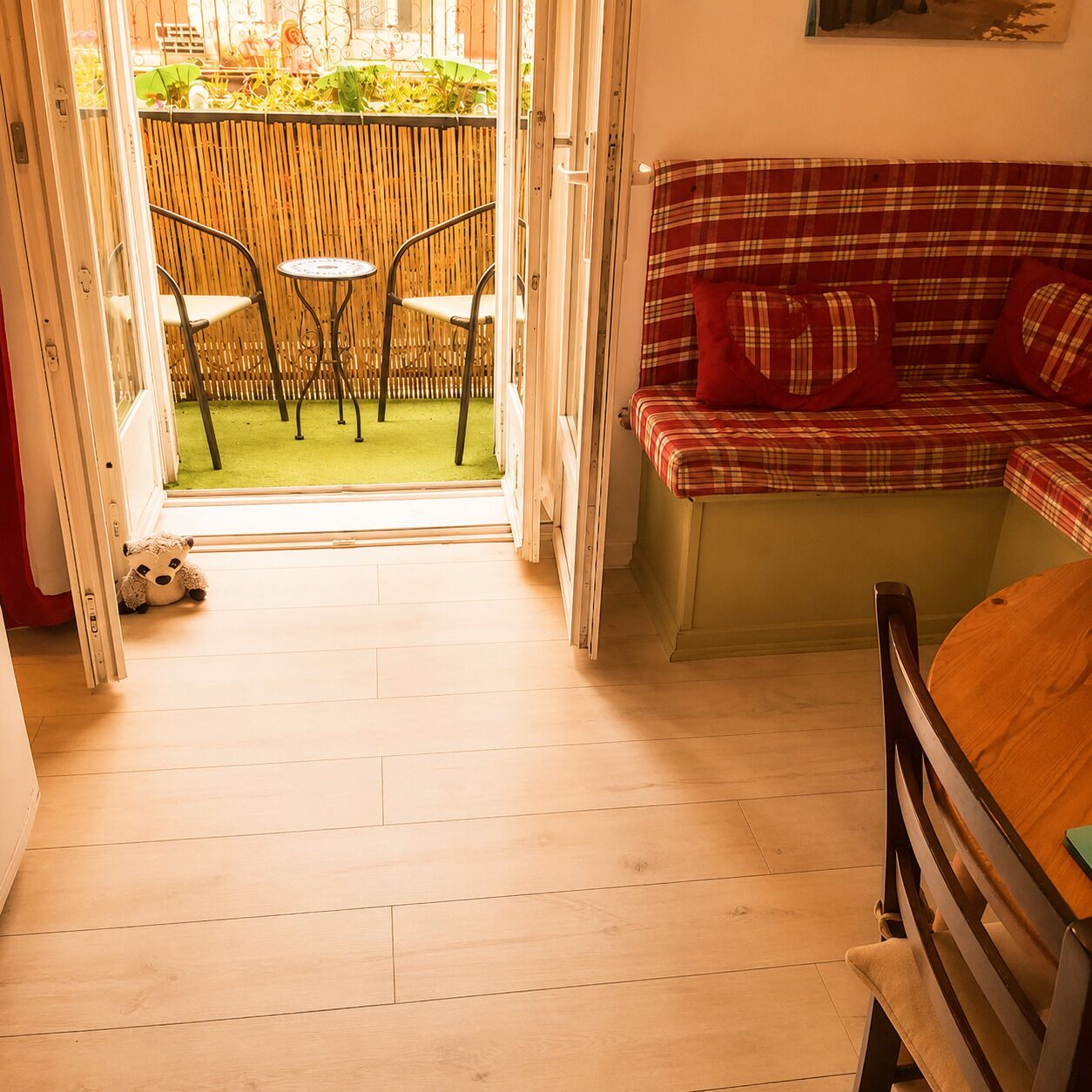 Een kamer met een houten vloer, een ruitjesbank met rode kussens, een klein rond tafeltje en twee stoelen.