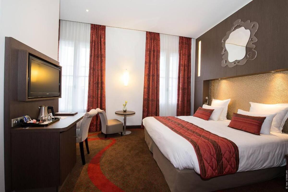 A hotel room with a bed, a desk with a coffee maker, a chair, a small table with a vase, and a wall-mounted TV.