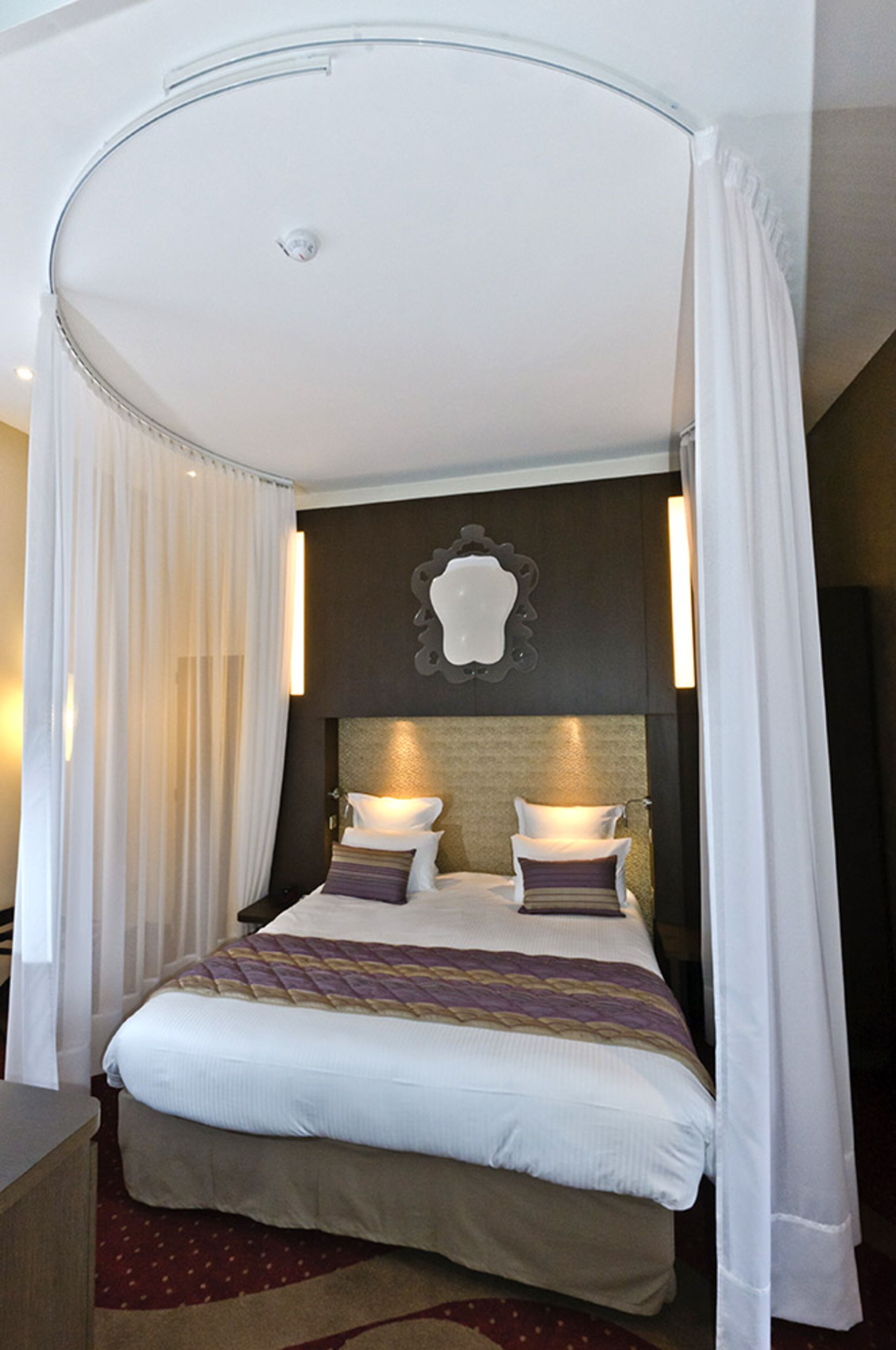 Une chambre d’hôtel avec un grand lit, des rideaux blancs et un grand miroir décoratif au mur.