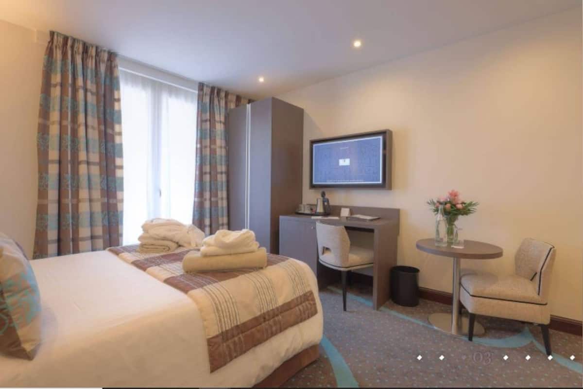 A hotel room with a bed, a desk, a chair, a television, and a small table with a vase of flowers.