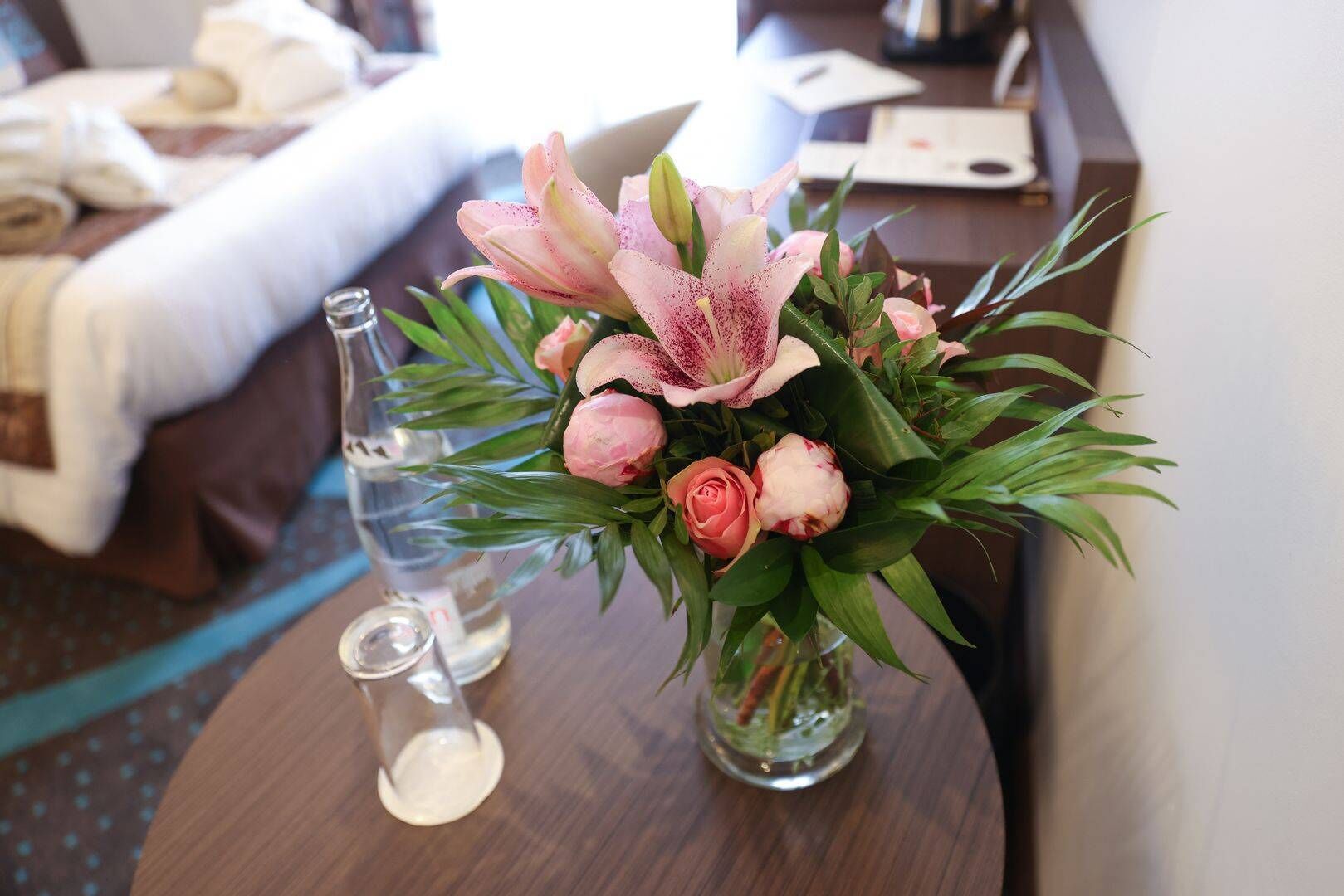 A hotel room with a bed, a small table with a vase of flowers, and a water bottle.