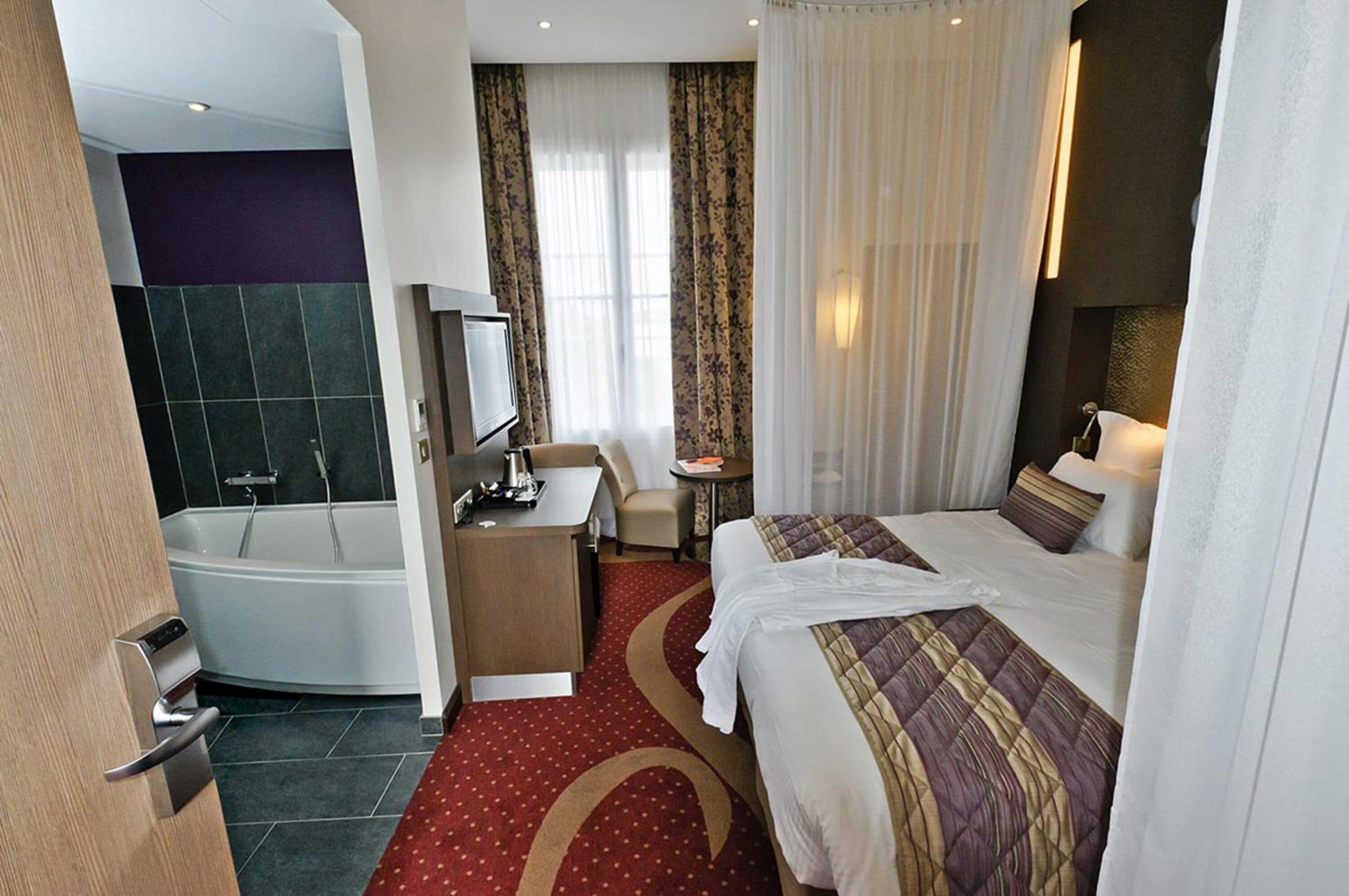 A hotel room with a large bed, a separate bathroom with a bathtub, a desk with a chair, and a small table with a lamp.