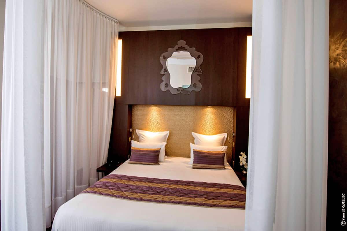 A hotel room with a large bed, two bedside tables, a headboard with a light fixture, and sheer curtains.