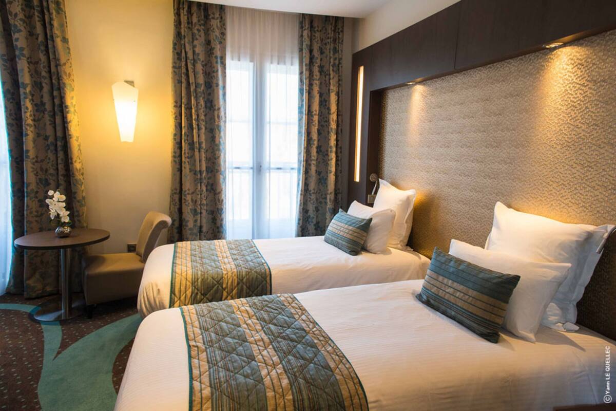 A hotel room with two beds, a small table, a chair, and a window with curtains.