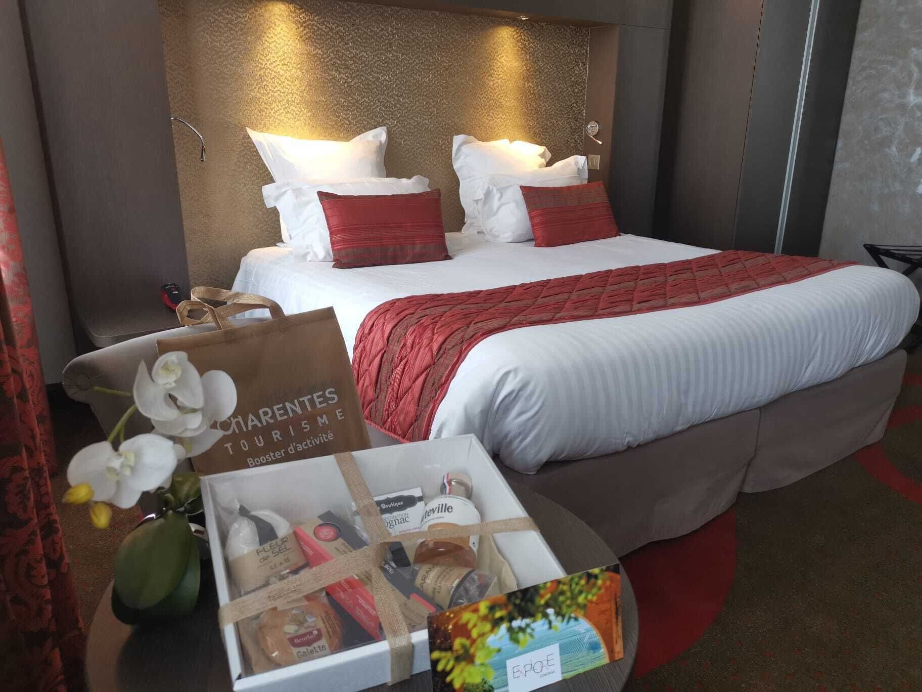 A hotel room with a large bed, two pillows, a red blanket, and a nightstand with a bag and a box of toiletries.