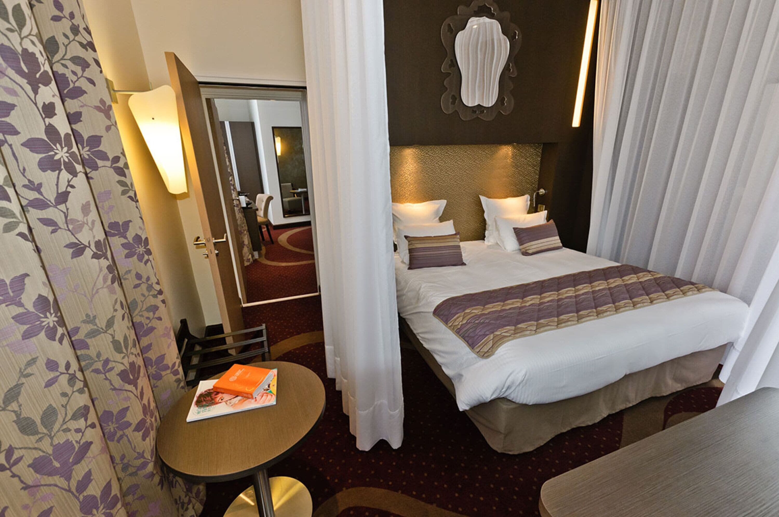 Une chambre d’hôtel avec un lit, une petite table sur laquelle se trouve un livre, un miroir et un luminaire fixé au mur.