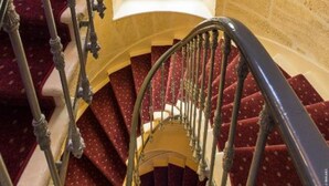 A spiral staircase with red carpeted steps and a metal railing, leading to a window with a view.