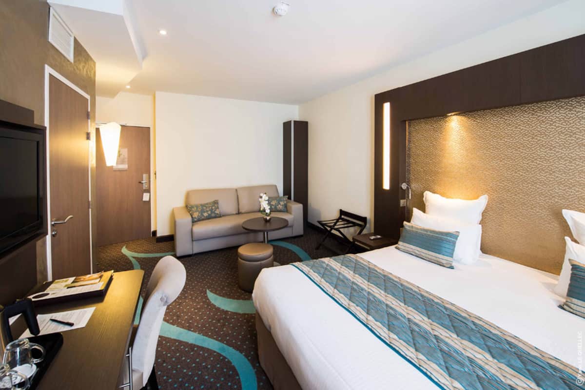 A hotel room with a bed, a sofa, a small table, a chair, and a desk with a computer and papers.