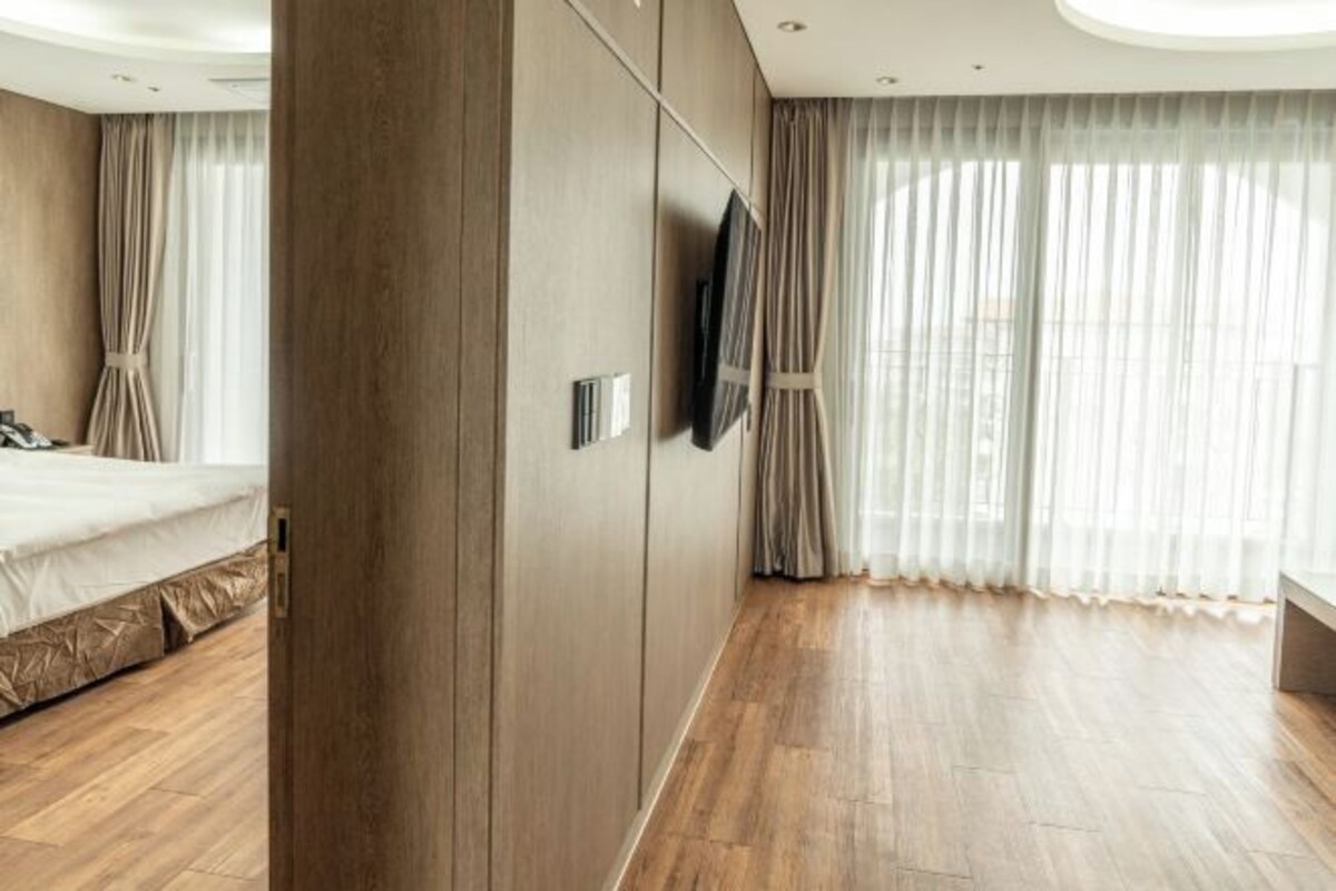 A modern hotel room with a wooden floor, a bed, a TV mounted on the wall, and large windows with sheer curtains.