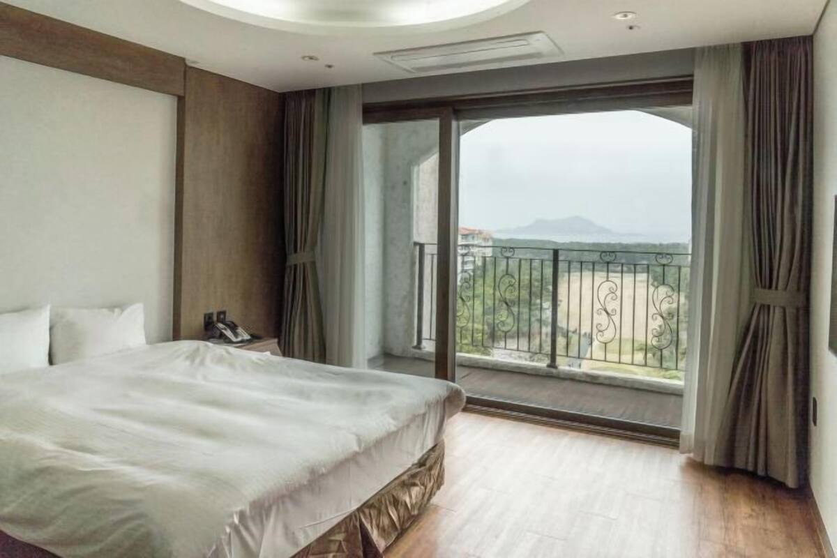 A hotel room with a large bed, a bedside telephone, a window with a view of the outdoors, and a balcony with a metal railing.