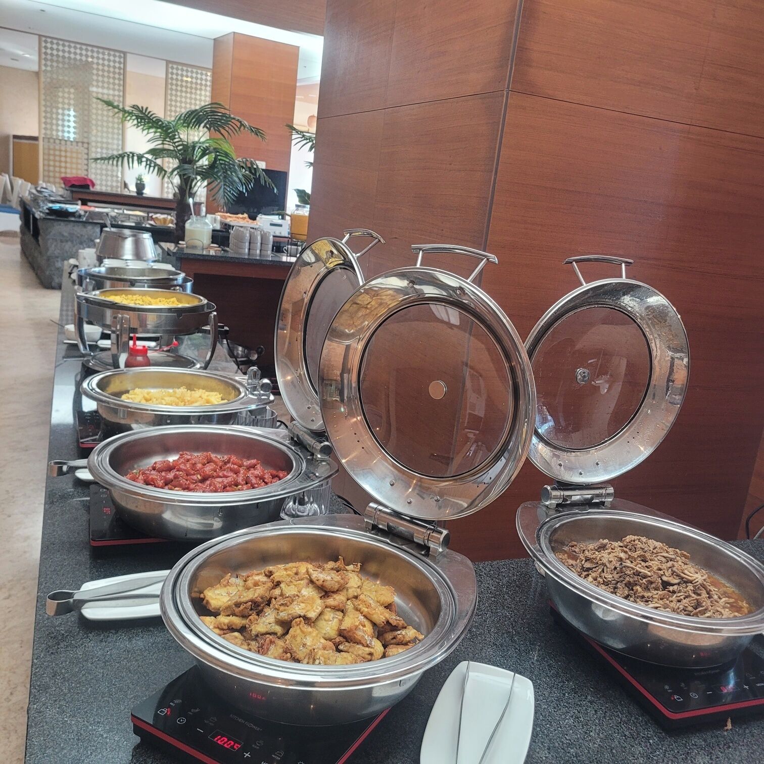 A buffet with various food items in metal containers, a coffee machine, and a plant in the background.