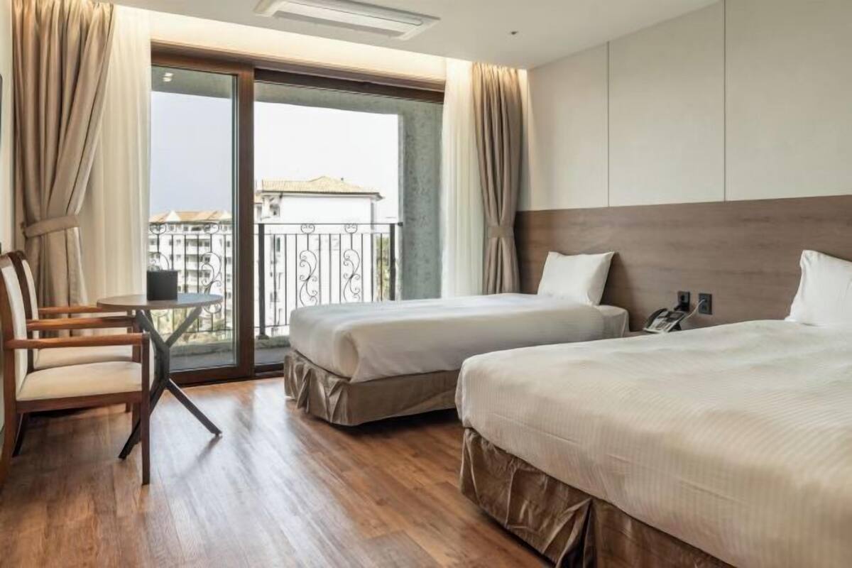 A hotel room with two beds, a small table, a chair, and a balcony with a view of buildings.