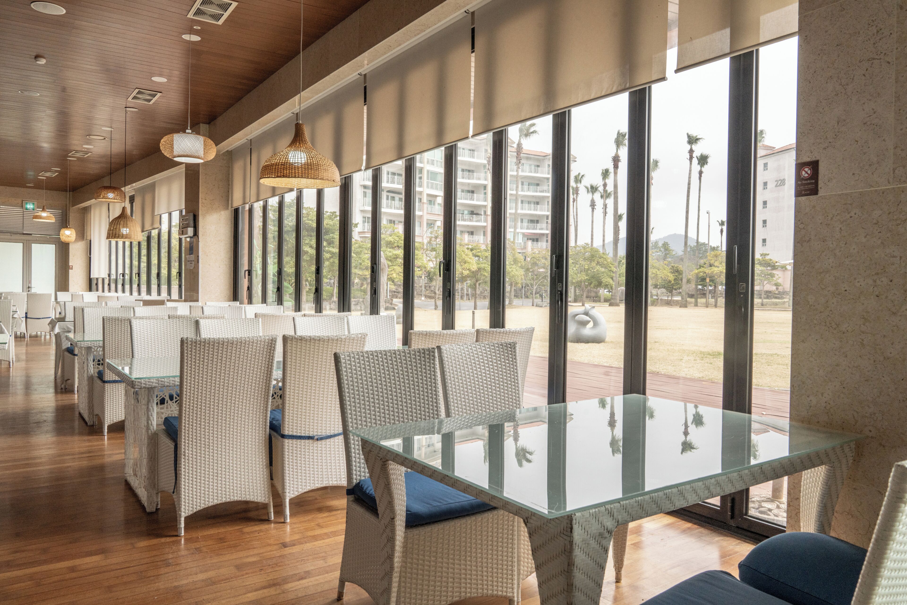 A modern dining area with wicker chairs, glass tables, and large windows offering a view of palm trees and buildings.