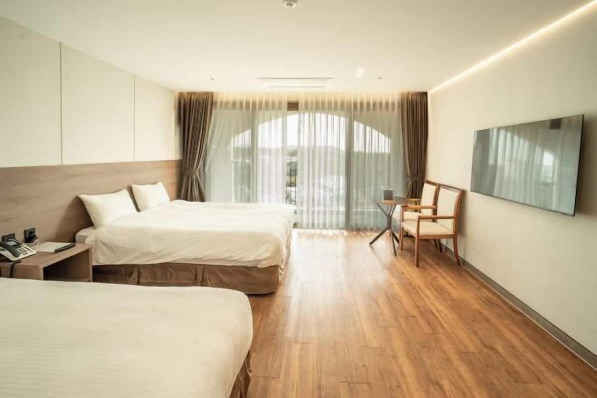 A hotel room with two beds, a desk, a chair, a TV, and a large window with curtains.