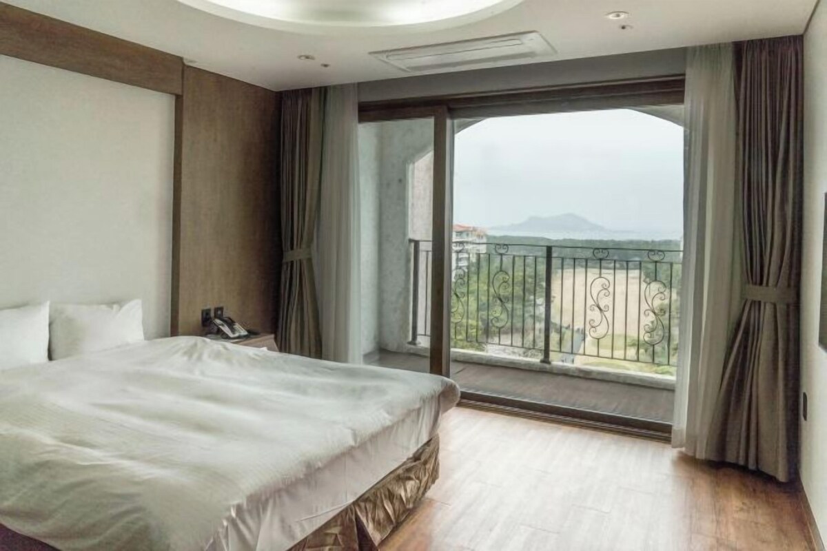 A hotel room with a large bed, a bedside telephone, a window with a view of the outdoors, and a balcony with a metal railing.