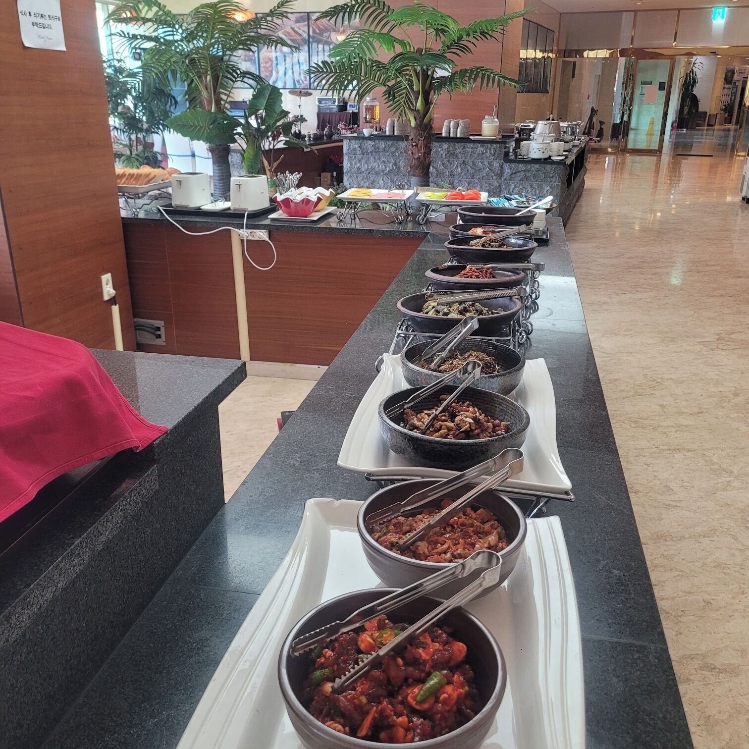 Een buffet met diverse gerechten in schalen, een receptiebalie en potplanten.