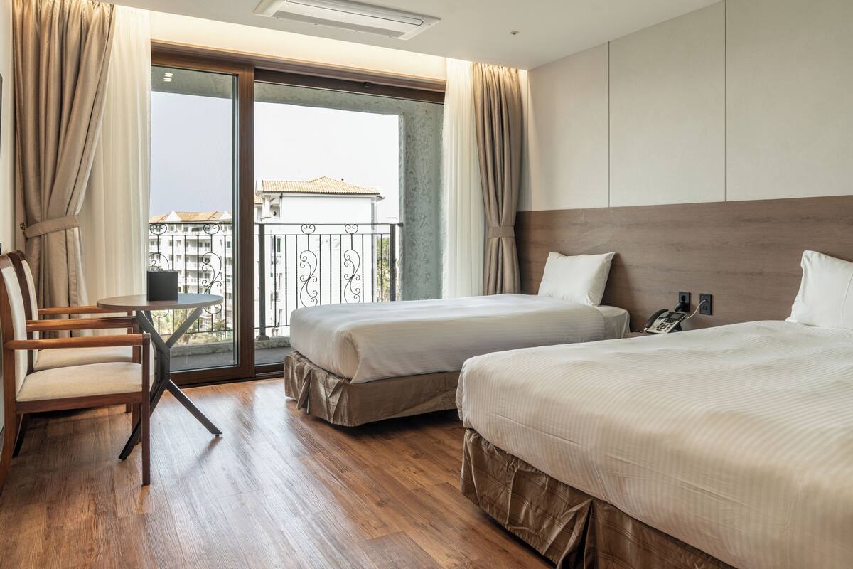 A hotel room with two beds, a small table, a chair, a balcony with a view, and a telephone on the wall.