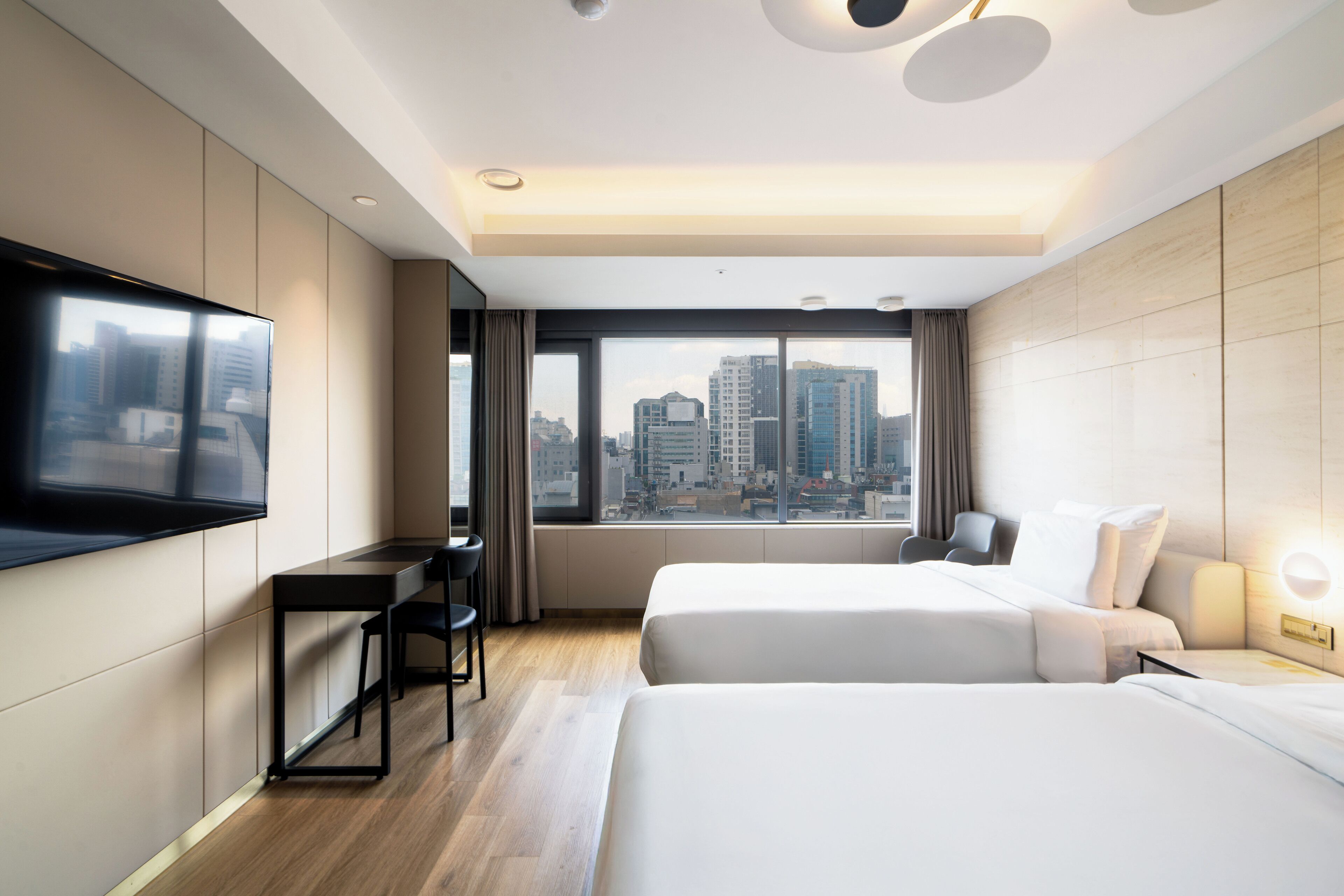 Habitación de hotel con dos camas, un escritorio, una silla, un televisor y un amplio ventanal con vista a la ciudad.