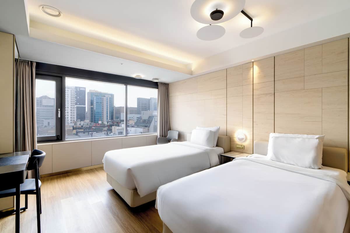 Habitación de hotel con dos camas, un escritorio y un ventanal grande con vistas a la ciudad.