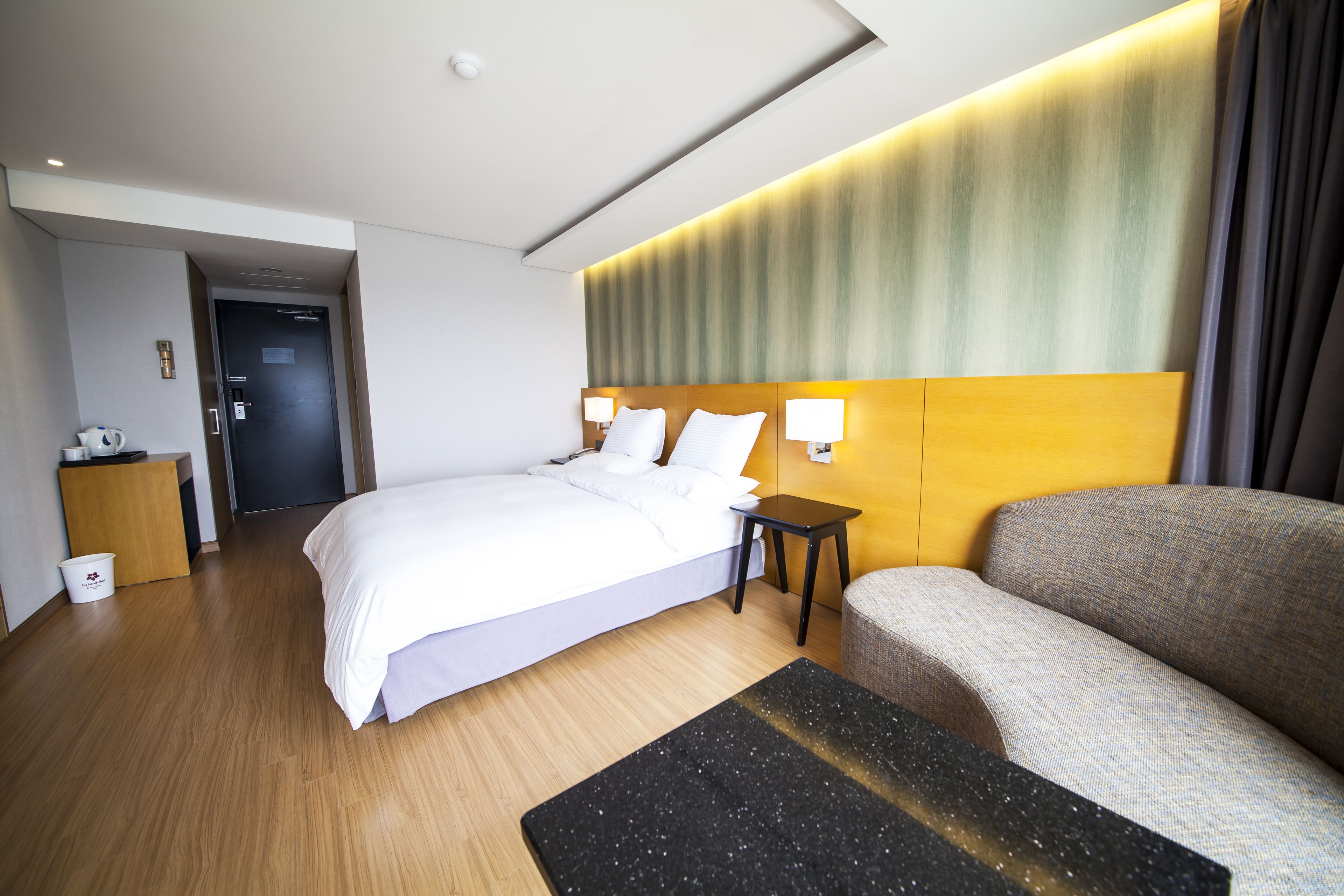 A modern hotel room with a bed, a sofa, a small table, and a wall-mounted lamp.