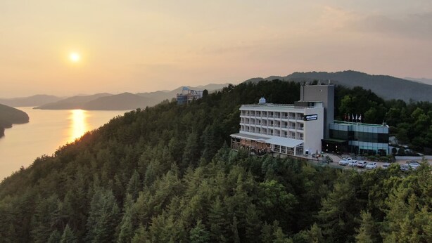 A hotel with multiple floors, surrounded by dense forest, overlooking a body of water during sunset. - Asia Lakeside Hotel (Jinju)