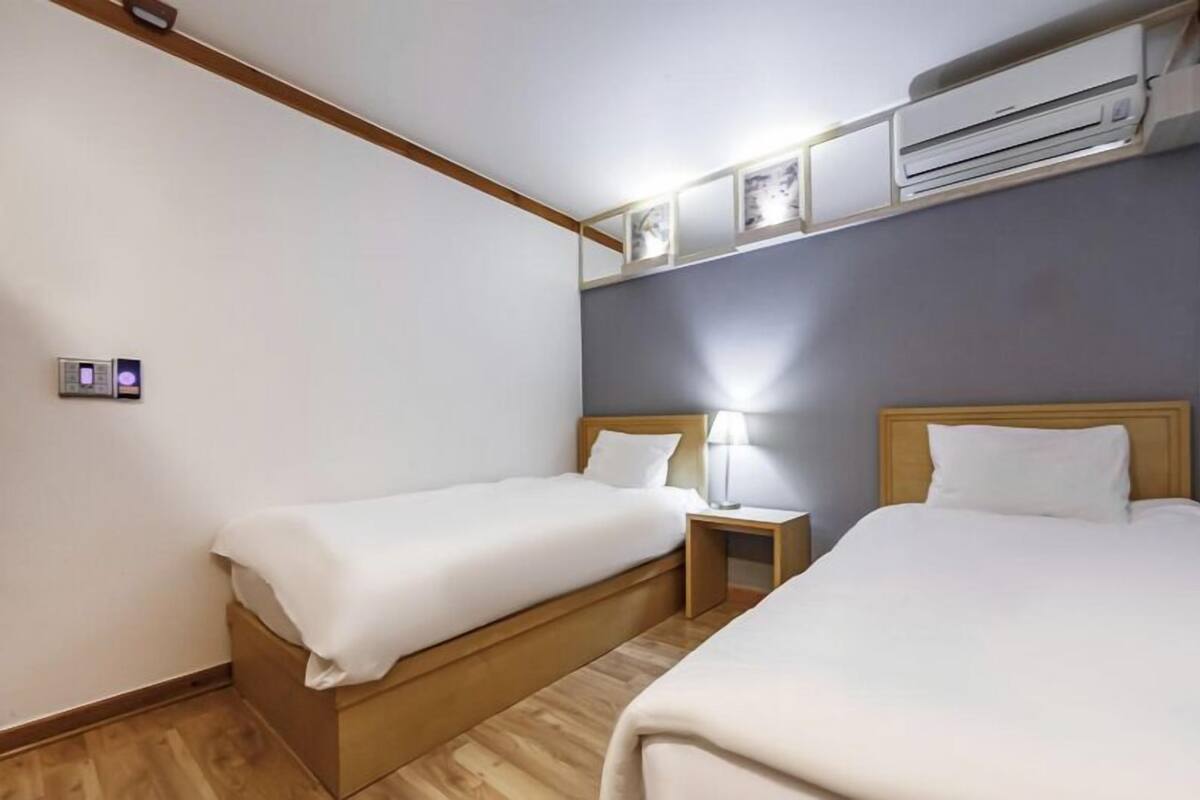 A hotel room with two single beds, a wooden floor, a wall-mounted air conditioner, and a bedside table with a lamp.