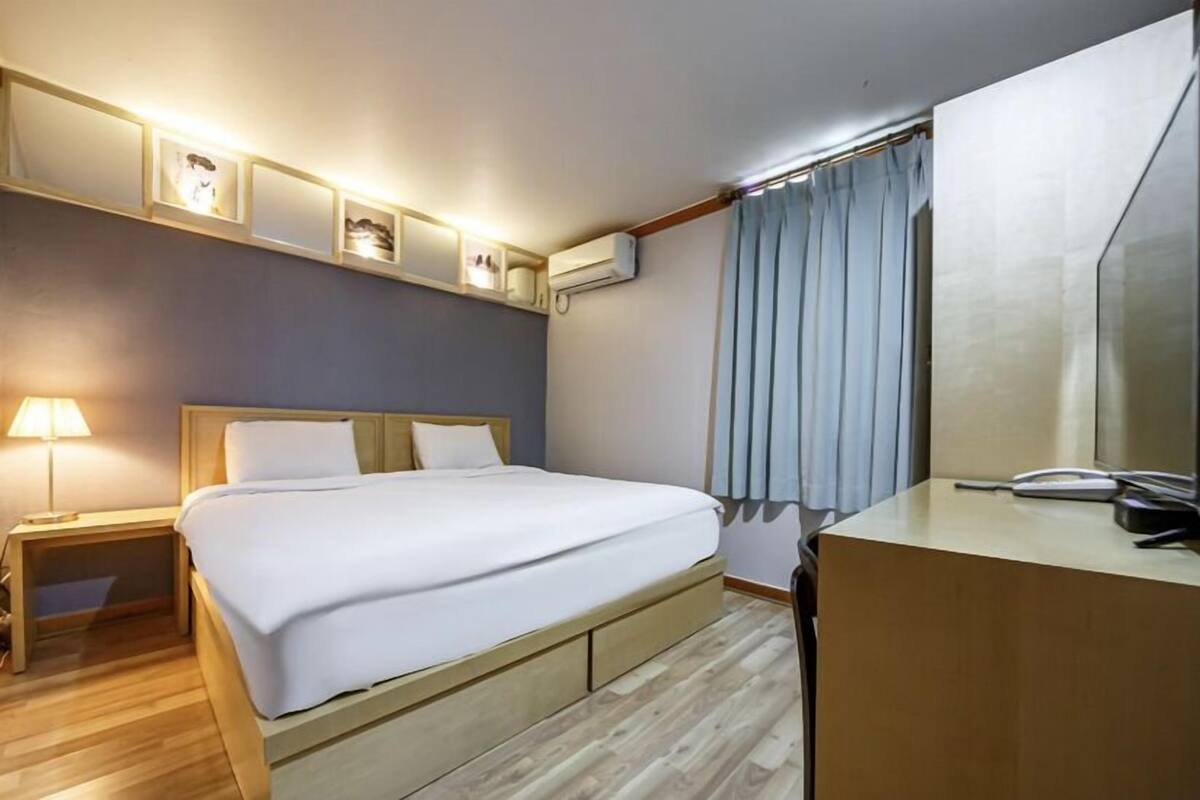 A hotel room with a bed, bedside table, lamp, and a desk with a telephone.