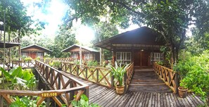 Een houten bruggetje dat leidt naar een bungalow met rieten dak, omgeven door weelderige begroeiing.