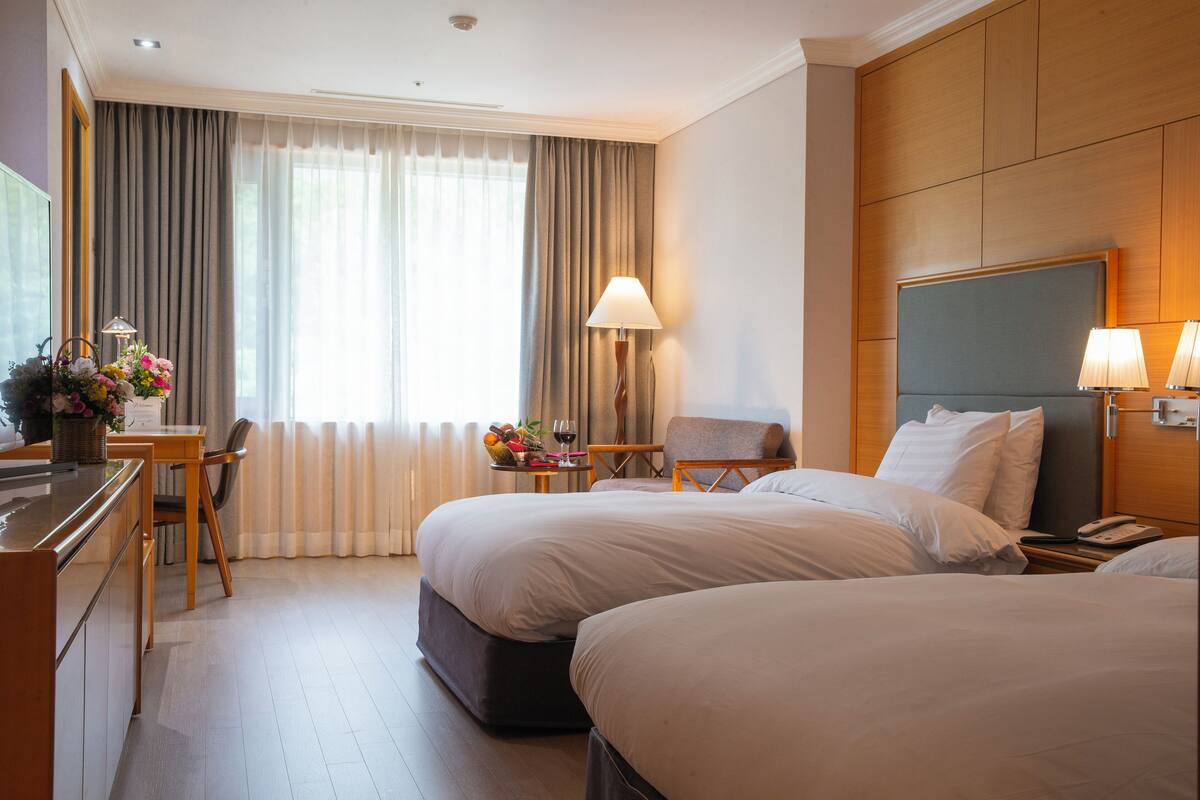 A hotel room with two beds, a desk with a lamp, a chair, and a window with curtains.